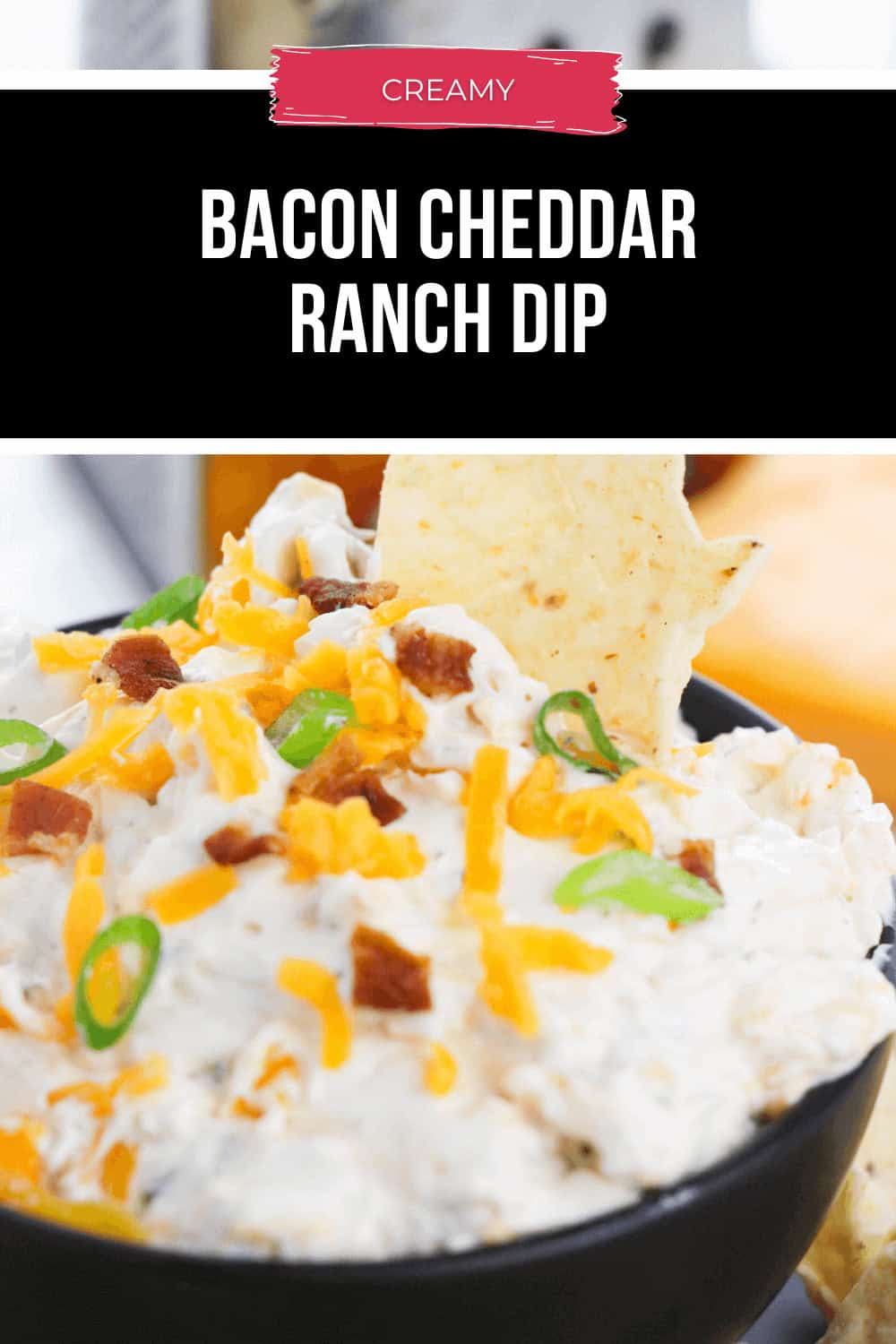 A bowl of Bacon Cheddar Ranch Dip, lusciously creamy and topped with shredded cheese, green onions, and crispy bacon bits, enticingly holds a tortilla chip for the perfect bite.