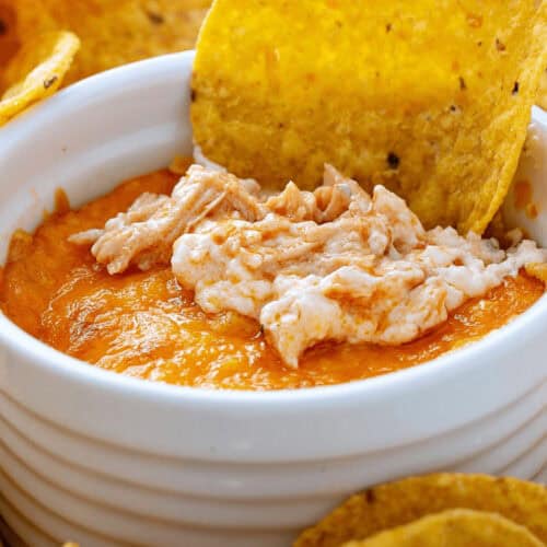 A bowl of orange and white Buffalo Chicken Wing Dip is surrounded by tortilla chips, with one chip partially dipped in the bowl.