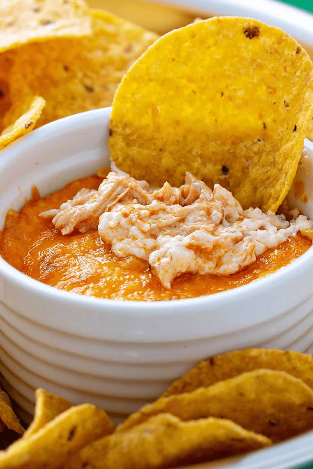 A bowl of orange and white Buffalo Chicken Wing Dip is surrounded by tortilla chips, with one chip partially dipped in the bowl.