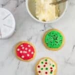 Three decorated sugar cookies and a bowl of creamy Buttercream Frosting for Cookies with a spoon on a marble surface. The cookies have red, green, and white icing with various sprinkles.