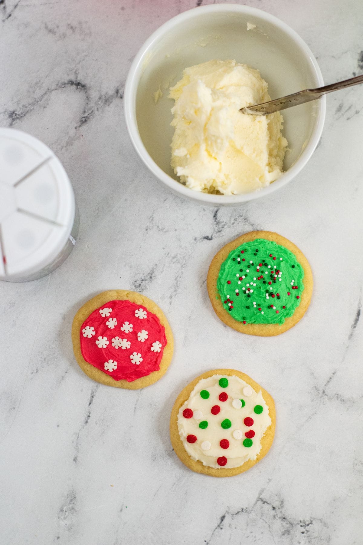 Three decorated sugar cookies and a bowl of creamy Buttercream Frosting for Cookies with a spoon on a marble surface. The cookies have red, green, and white icing with various sprinkles.