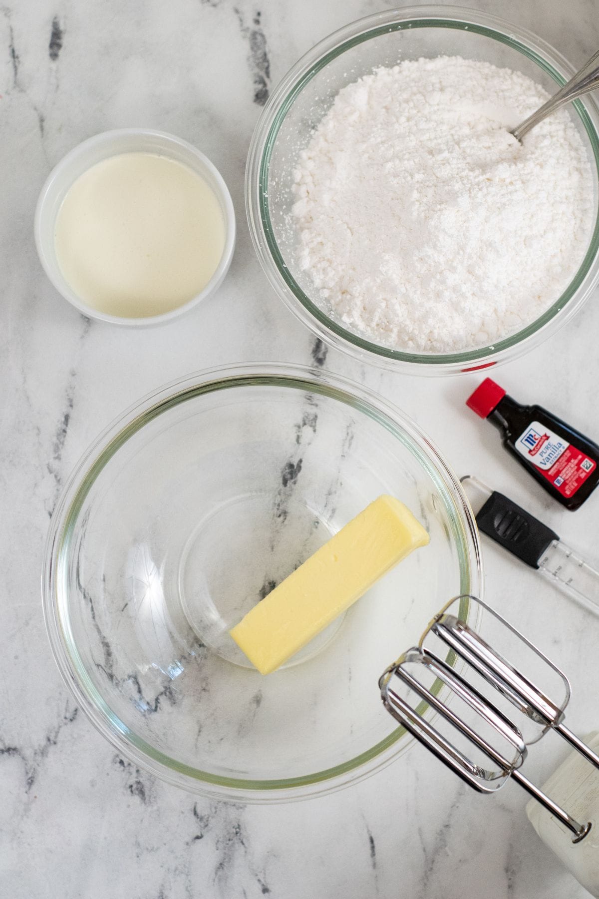 Glass bowls with powdered sugar, cream, vanilla extract, and a stick of butter are arranged on a marble countertop.