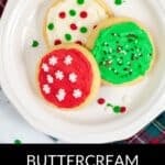 Three sugar cookies with red, green, and white buttercream frosting for cookies and colorful sprinkles sit on a white plate next to a plaid cloth.
