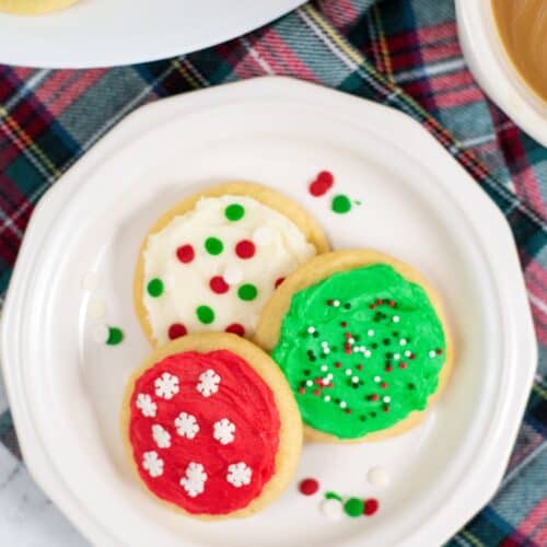 Three sugar cookies with red, green, and white buttercream frosting for cookies and sprinkles sit on a white plate, placed on a plaid cloth next to a cup of coffee.