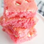 Three pink Cake Mix Strawberry Brownies with white chocolate chips are stacked on a white plate, with a striped napkin in the background.