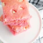 Three pink desserts with white chocolate chips are stacked on a white plate, with strawberries and a checkered cloth in the background.