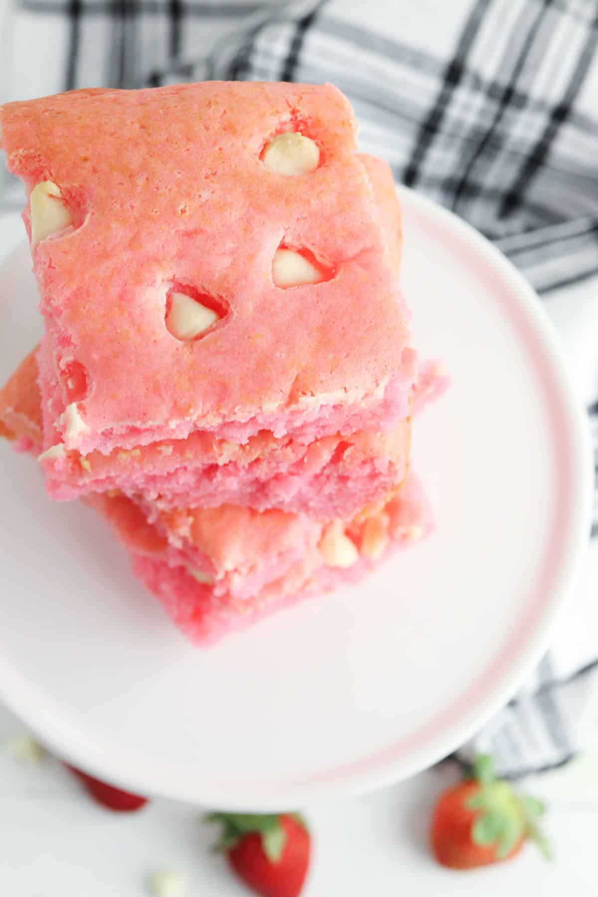 Three pink desserts with white chocolate chips are stacked on a white plate, with strawberries and a checkered cloth in the background.