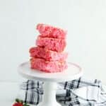Four Cake Mix Strawberry Brownies stacked on a white cake stand, surrounded by fresh strawberries and a black-and-white striped cloth on a white background.