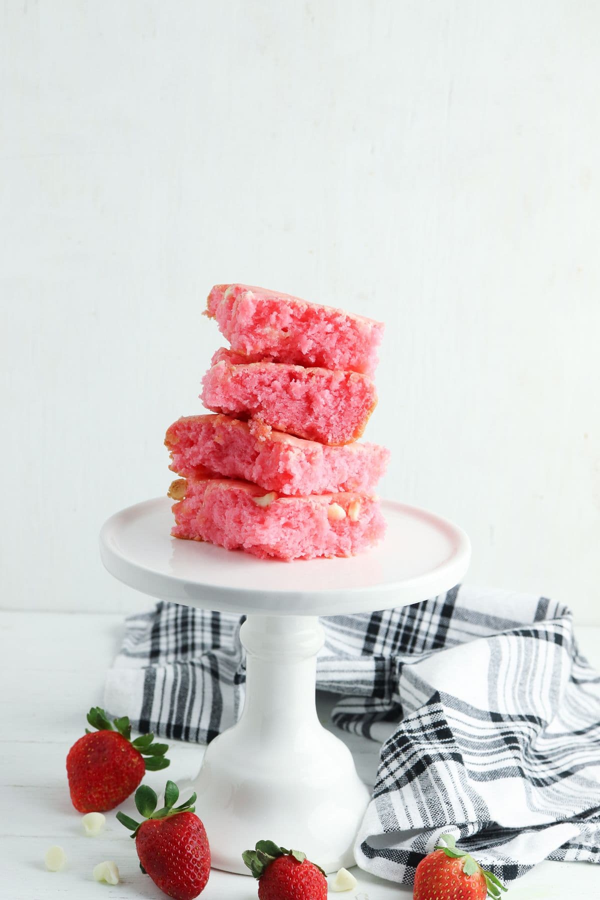 Four Cake Mix Strawberry Brownies stacked on a white cake stand, surrounded by fresh strawberries and a black-and-white striped cloth on a white background.