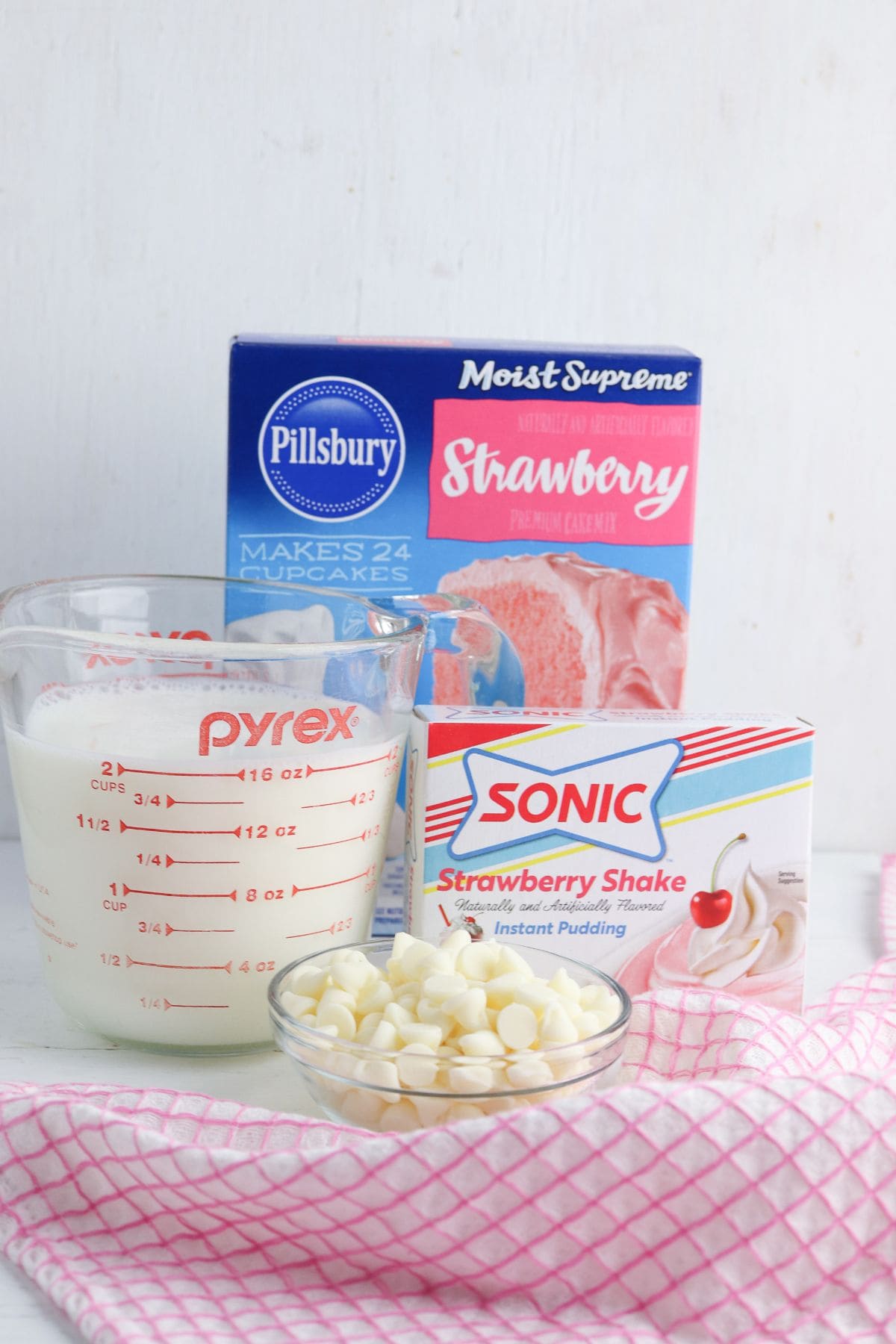 A box of Mix,  instant pudding, a measuring cup of milk, white chocolate chips in a bowl, and a pink dish towel on a white surface.