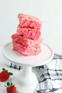 Four pink Cake Mix Strawberry Brownies stacked on a white cake stand, with fresh strawberries and a black-and-white towel nearby.
