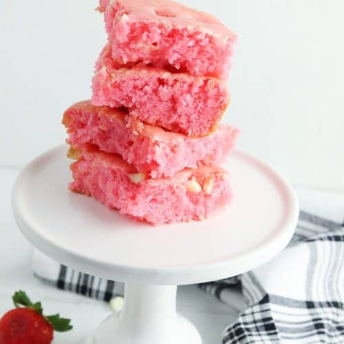 Four pink Cake Mix Strawberry Brownies stacked on a white cake stand, with fresh strawberries and a black-and-white towel nearby.