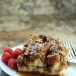 Two pieces of Cinnamon Roll French Toast bread pudding with streusel topping, drizzled with syrup on a white plate, served with fresh raspberries and a fork.