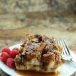 A plate with two pieces of Cinnamon Roll French Toast casserole topped with syrup, served with a side of fresh raspberries and a fork.
