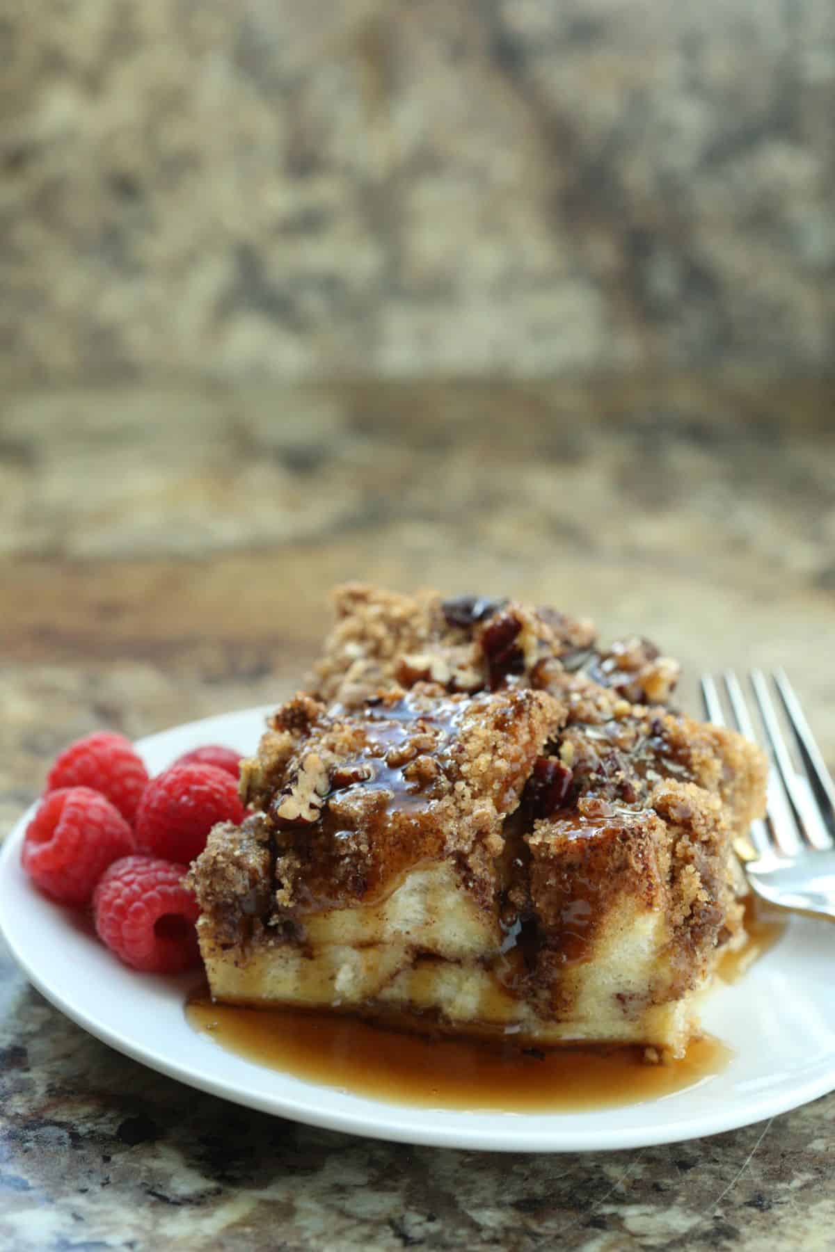 A plate with two pieces of casserole drizzled with syrup and topped with pecans, served alongside fresh raspberries and a fork.