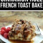 A serving of Cinnamon Roll French Toast bake with syrup and fresh raspberries on a white plate, next to a fork, rests on a marbled surface.