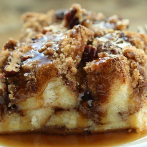A close-up of a slice of bread pudding inspired by Cinnamon Roll French Toast, topped with pecans, crumbly streusel, and drizzled with syrup on a white plate.