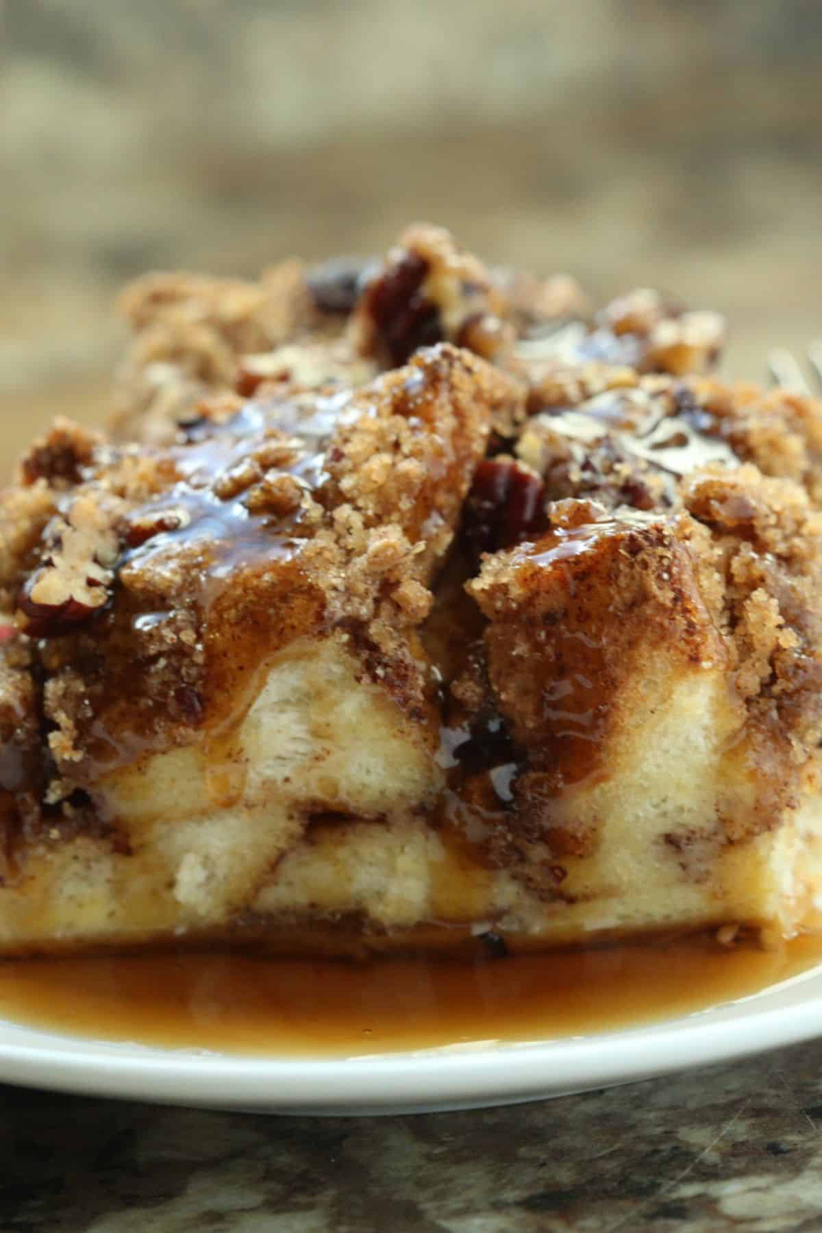 A close-up of a slice of bread pudding inspired by Cinnamon Roll French Toast, topped with pecans, crumbly streusel, and drizzled with syrup on a white plate.
