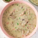 A pink bowl filled with creamy chicken mushroom soup containing sliced mushrooms, herbs, and vegetables; fresh chopped herbs and bread are nearby.