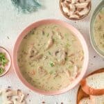 A bowl of Creamy Chicken Mushroom Soup with sliced mushrooms and herbs, surrounded by bread slices, spices, and fresh mushroom pieces on a light surface.