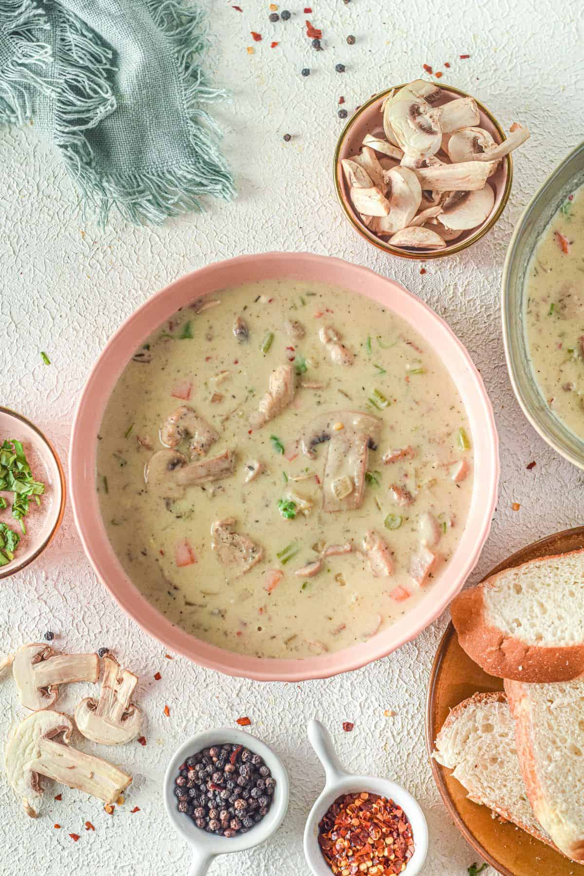 A bowl of Creamy Chicken Mushroom Soup with sliced mushrooms and herbs, surrounded by bread slices, spices, and fresh mushroom pieces on a light surface.
