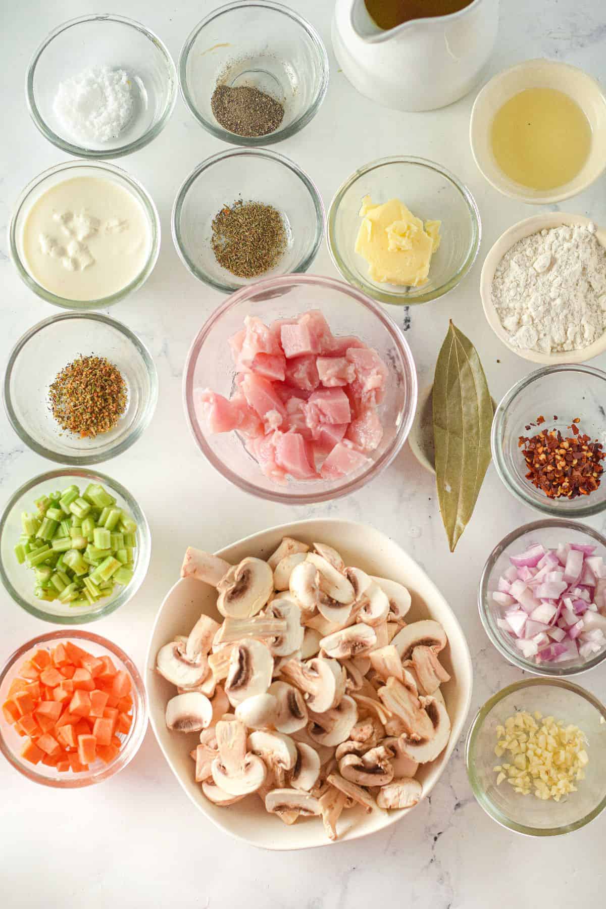 Several glass bowls containing chopped vegetables, raw chicken, sliced mushrooms, spices, and liquids are arranged on a white surface for preparing Creamy Chicken Mushroom Soup.