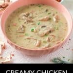 A bowl of Creamy Chicken Mushroom Soup with tender chicken and vegetables, garnished with herbs, sits on a light surface near bread and red pepper flakes.