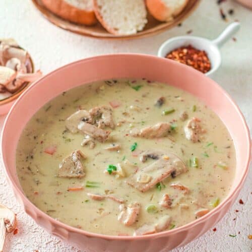 A bowl of Creamy Chicken Mushroom Soup with chunks of meat and vegetables, served alongside slices of bread and a dish of red pepper flakes in the background.