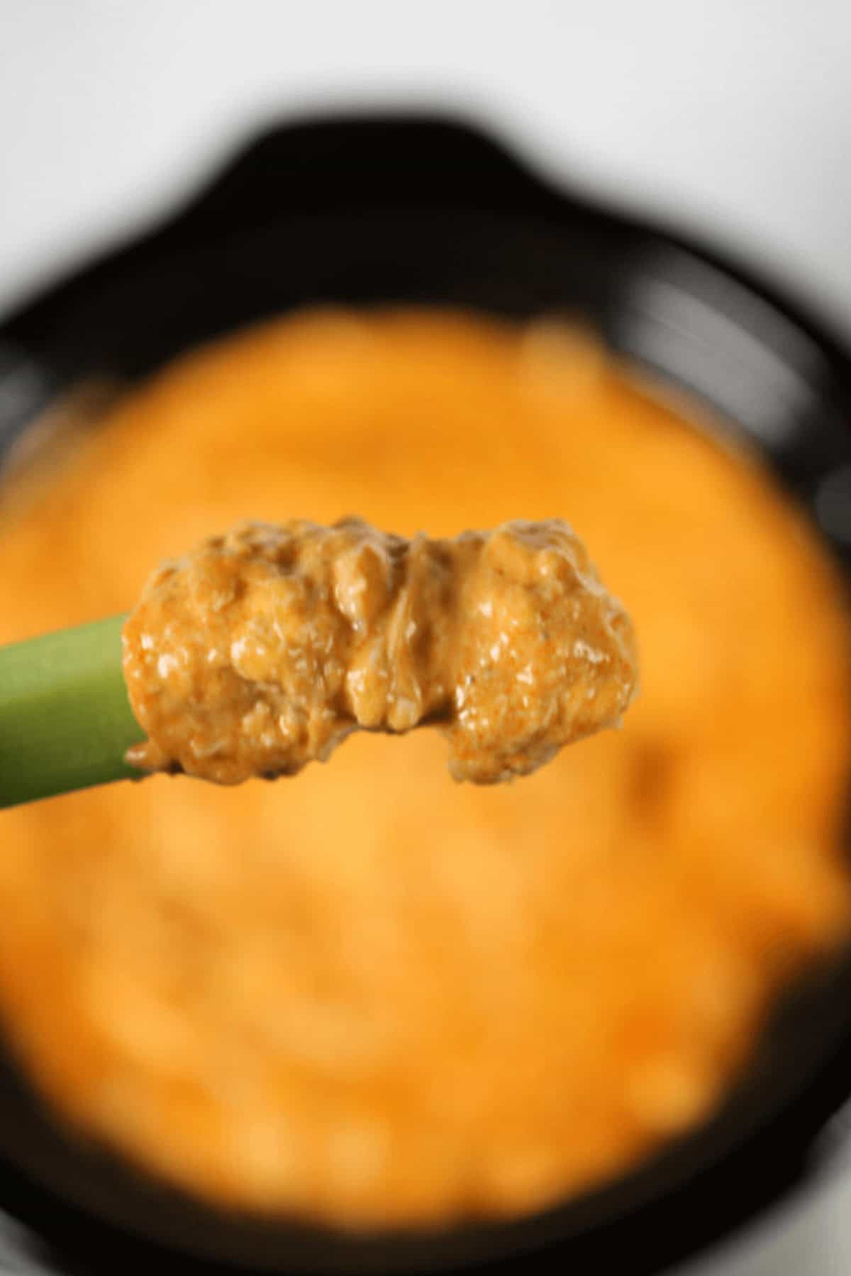 A close-up of a celery stick dipped, held above a dish brimming with more of the same tantalizing dip.
