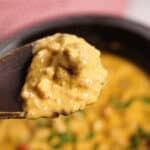 Close-up of a wooden spoon holding curry with visible chunks of meat and vegetables, above a bowl of the same dish. Reminiscent of a Crock Pot creation, the dish is creamy and garnished with fresh green herbs, offering a hearty warmth akin to a comforting cheeseburger dip.