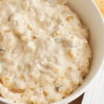 A white bowl brimming with creamy cheese corn dip features visible bits of corn and black olives, all resting invitingly on a wooden surface.