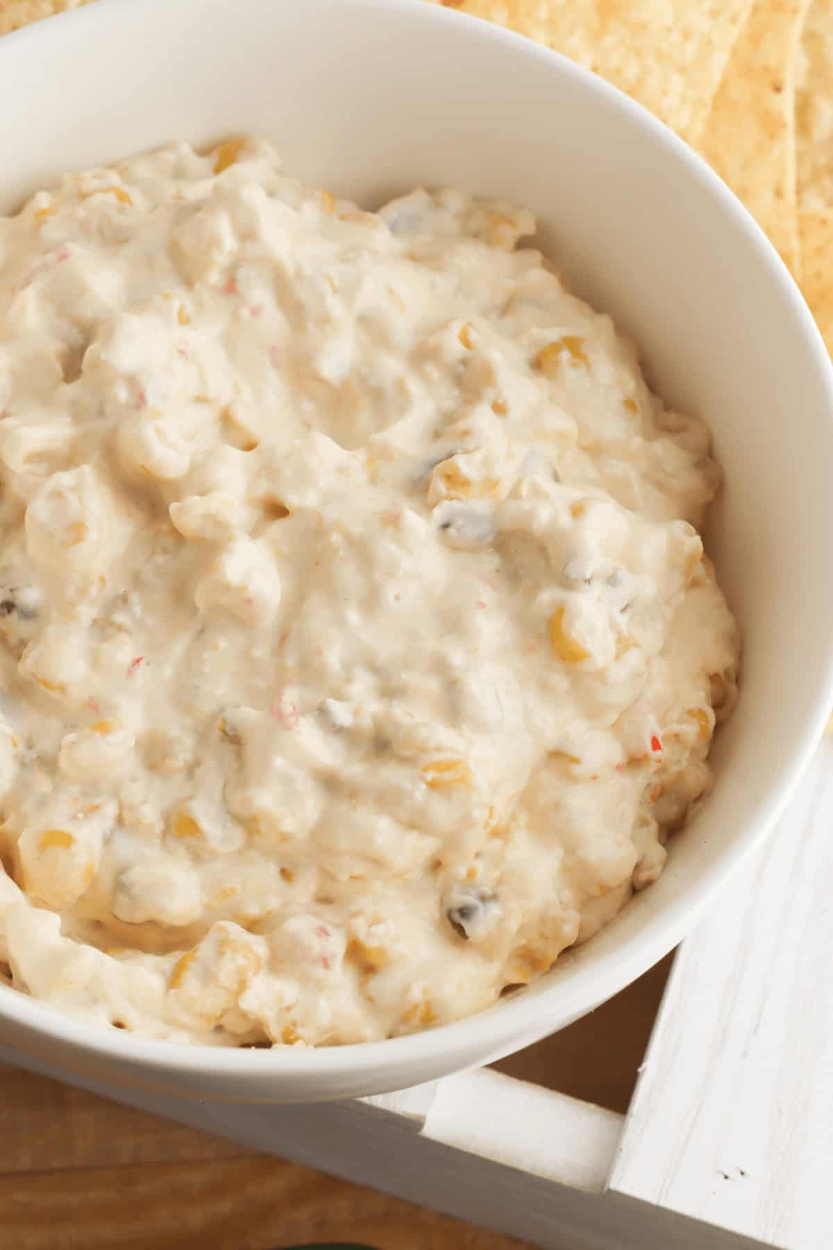A white bowl brimming with creamy cheese corn dip features visible bits of corn and black olives, all resting invitingly on a wooden surface.