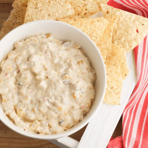 A bowl of creamy cream cheese corn dip is surrounded by tortilla chips on a white tray. Two jalapeños and a red-striped cloth are nearby, adding a splash of color and flavor to this inviting snack scene.
