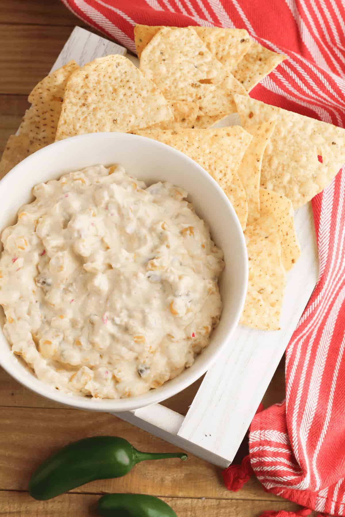 A bowl of creamy cream cheese corn dip is surrounded by tortilla chips on a white tray. Two jalapeños and a red-striped cloth are nearby, adding a splash of color and flavor to this inviting snack scene.