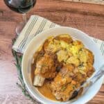 A bowl of Instant Pot Beef Short Ribs with sauce, roasted potatoes, and herbs sits next to a glass of red wine on a wooden table with a striped napkin.