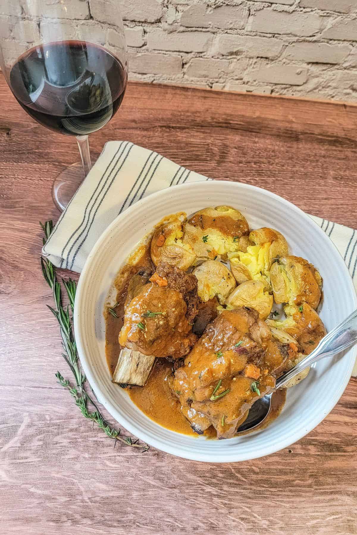 A bowl of Instant Pot Beef Short Ribs with sauce, roasted potatoes, and herbs sits next to a glass of red wine on a wooden table with a striped napkin.