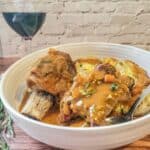 A bowl of Instant Pot Beef Short Ribs in sauce served with roasted potatoes, garnished with herbs, next to a glass of red wine on a wooden table.
