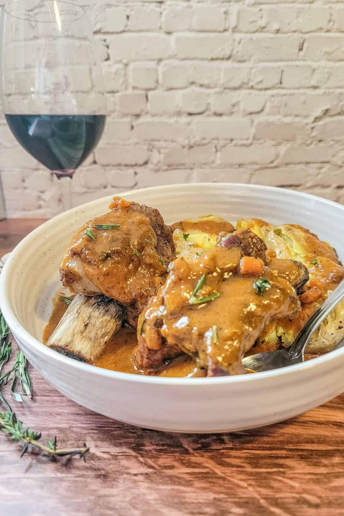 A bowl of Instant Pot Beef Short Ribs in sauce served with roasted potatoes, garnished with herbs, next to a glass of red wine on a wooden table.