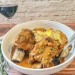 A bowl of Instant Pot Beef Short Ribs with rich gravy, served over roasted potatoes, sits on a wooden table beside a sprig of rosemary and a glass of red wine.