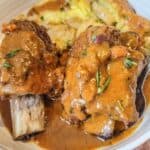 A close up on the meat garnished with fresh herbs, served alongside smashed potatoes in a white bowl.