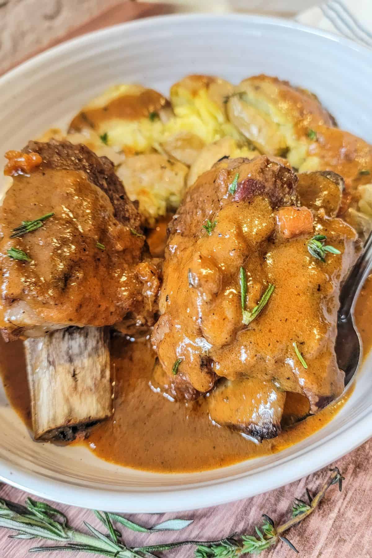 A close up on the meat garnished with fresh herbs, served alongside smashed potatoes in a white bowl.