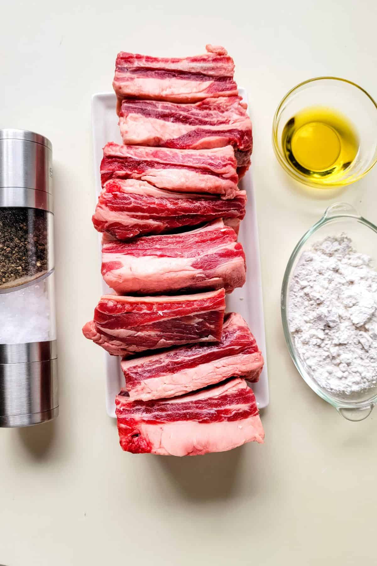 Rectangular slices of raw meat on a white plate, with bowls of olive oil and flour, plus a salt and pepper grinder on a white surface.