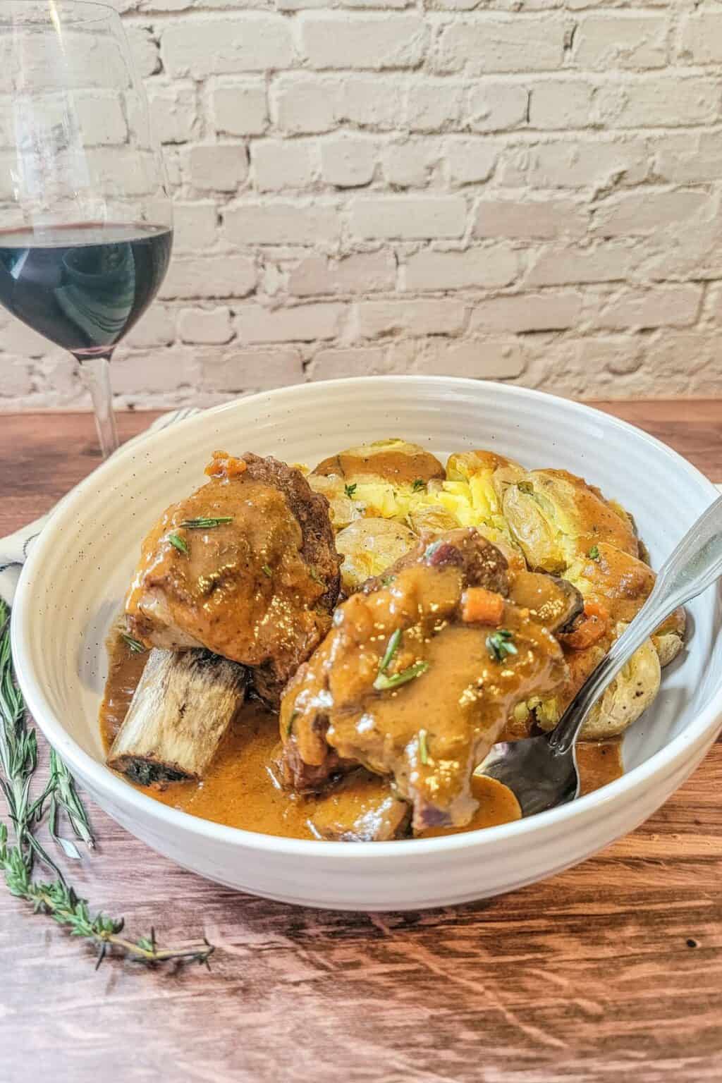 A bowl of tender Instant Pot Beef Short Ribs with rich gravy, served over sliced potatoes, garnished with rosemary, and paired with a glass of red wine on a rustic wooden table.