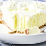 A slice of creamy Lemonade Frozen Pie with a graham cracker crust is served on a white plate. A fork with a piece of the pie lies on the plate, and the pie is topped with whipped cream.