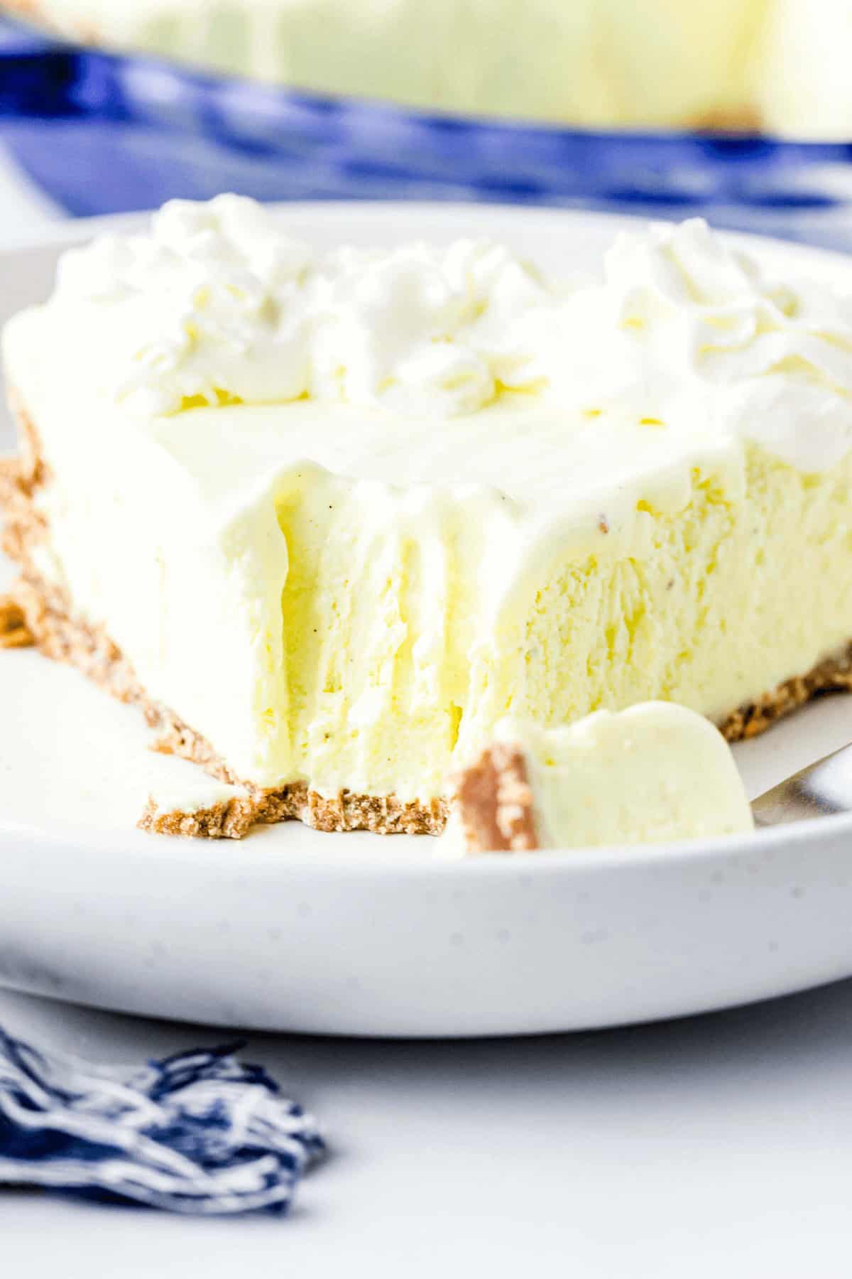 A slice of creamy Lemonade Frozen Pie with a graham cracker crust is served on a white plate. A fork with a piece of the pie lies on the plate, and the pie is topped with whipped cream.