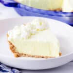 A slice of creamy dessert with a graham cracker crust and whipped cream topping on a white plate, with the rest of the pie in a blue dish in the background.