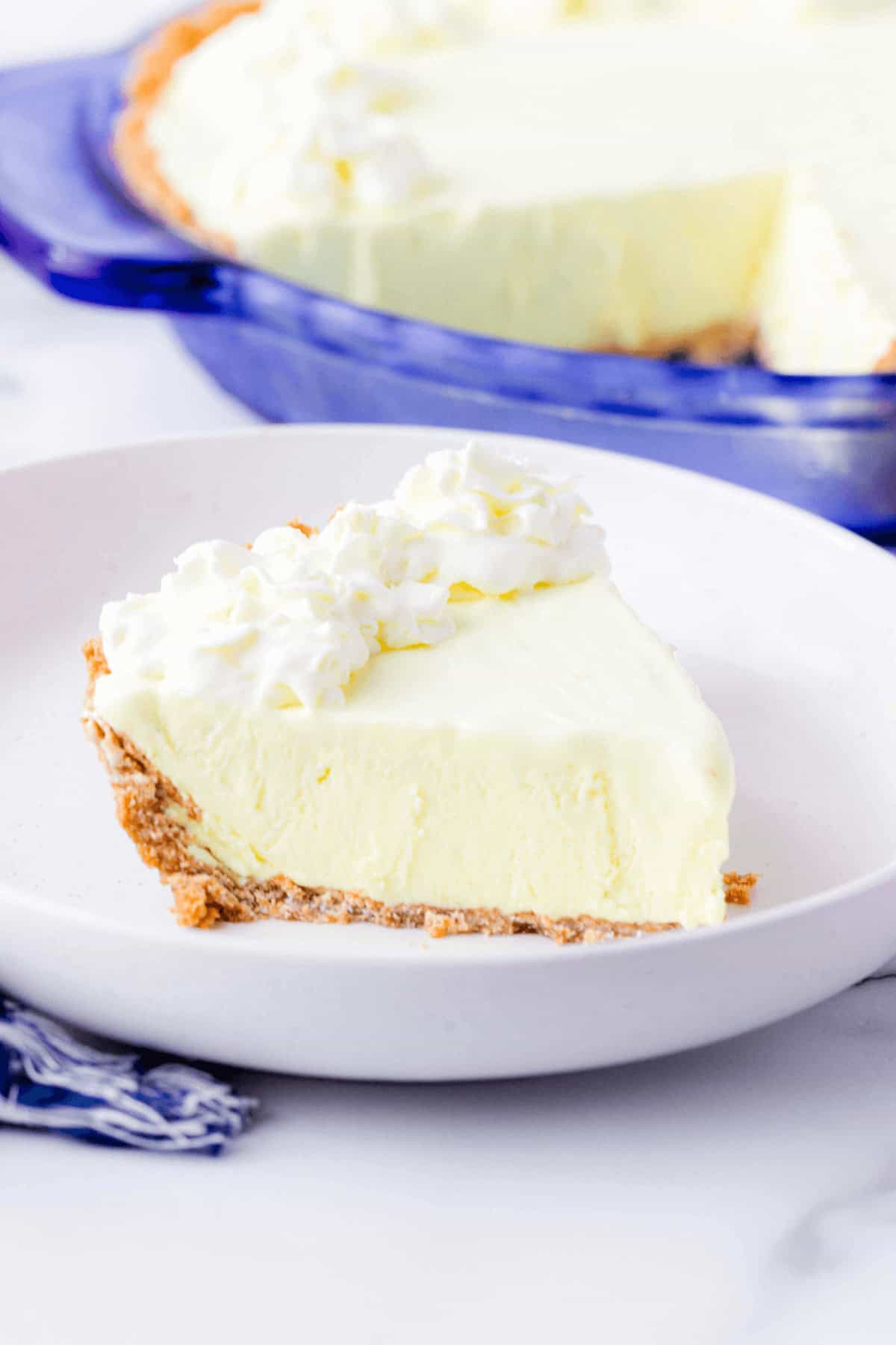 A slice of creamy dessert with a graham cracker crust and whipped cream topping on a white plate, with the rest of the pie in a blue dish in the background.