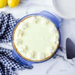 A lemonade frozen pie with a graham cracker crust and whipped cream dollops rests in a blue pie dish, placed next to two lemons, a checkered cloth, and stacked white plates on a marble surface.