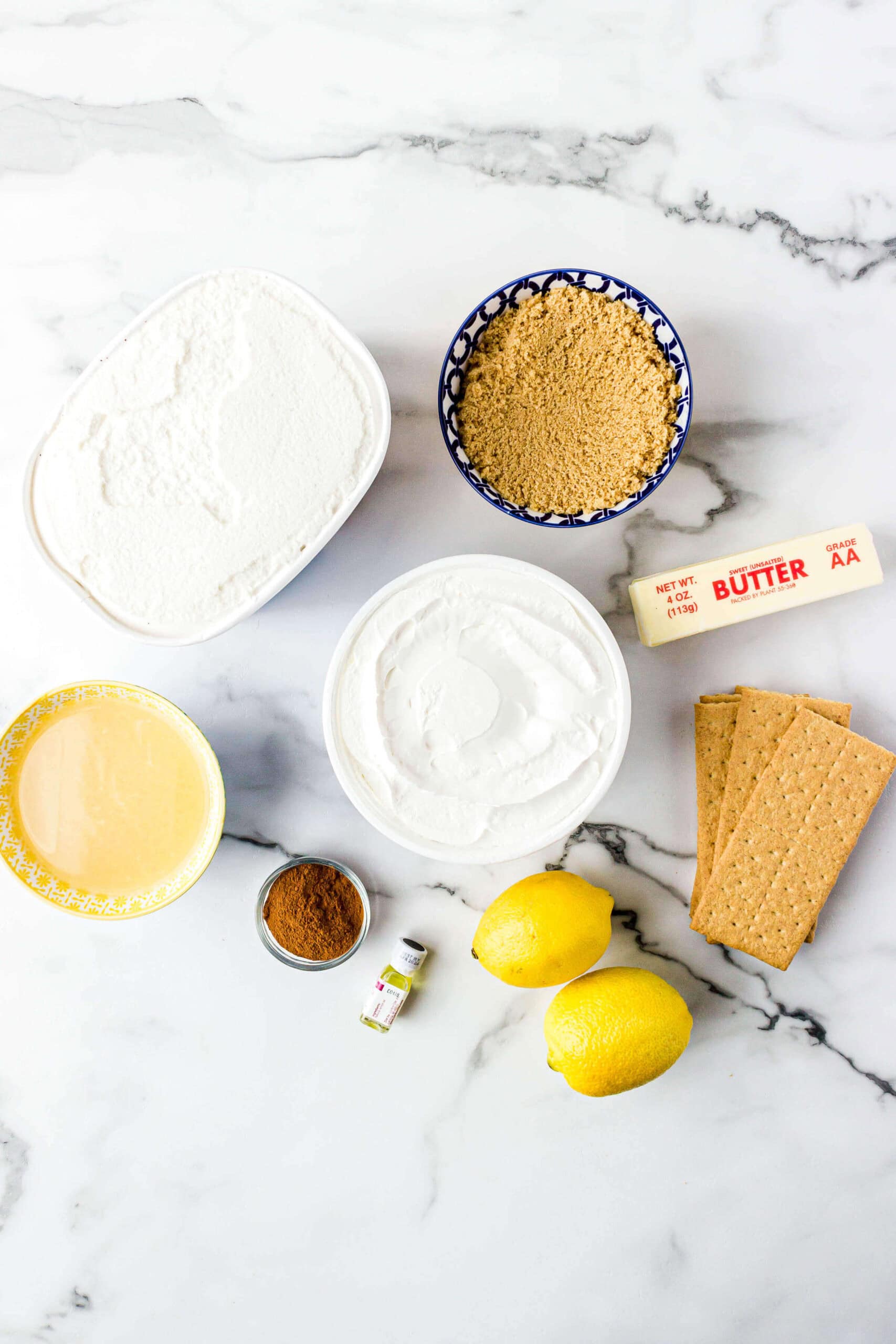 Ingredients for a Lemonade Frozen Pie recipe arranged on a marble surface, including flour, brown sugar, butter, graham crackers, lemons, whipped cream, cinnamon, and vanilla extract.