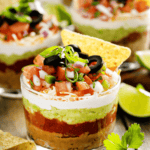 Glass cup showcasing a vibrant 5 Layer Dip with beans, guacamole, sour cream, salsa, and cheese. It's topped artistically with tomatoes, olives, and green onions. A tortilla chip perches invitingly on the rim.